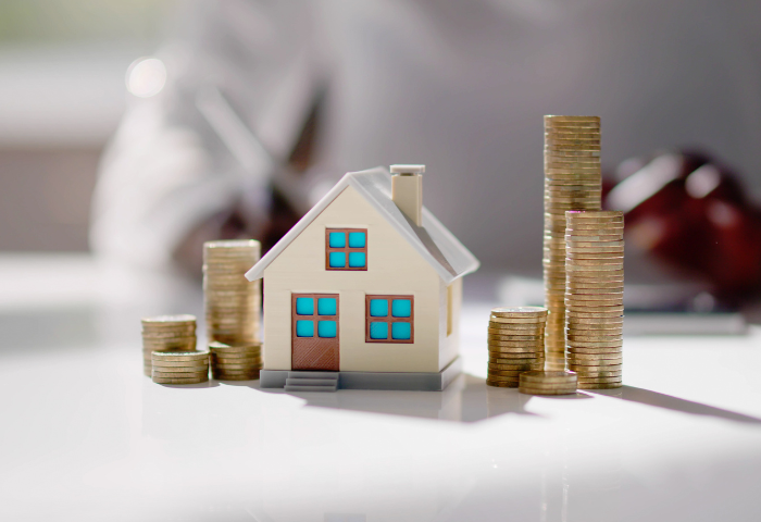 A small model of a house rests on a table. Several stacks of pound coins surround the two sides of the model.