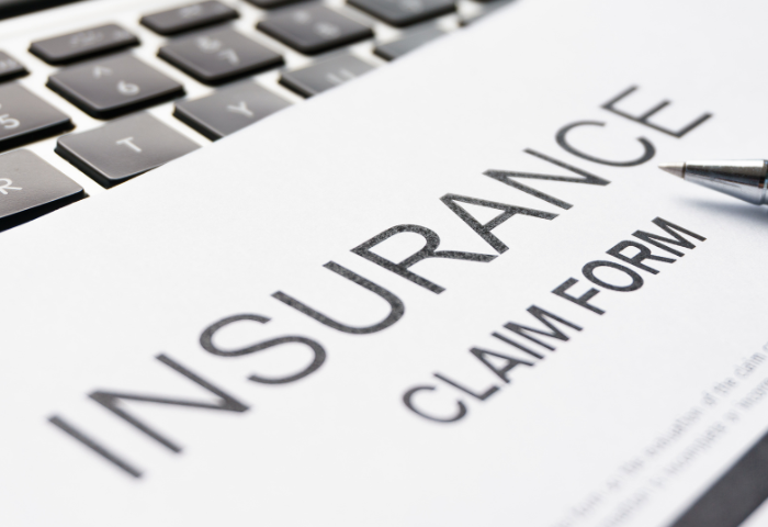 An extreme close up of the words "Insurance Claim Form" written in capitals on a piece of paper. The tip of a pen is resting on the paper and a laptop keyboard can be seen in the background.